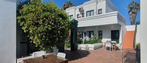 Un patio con una mesa de madera y sillas blancas, un patio de ladrillo y un edificio blanco con una puerta verde.