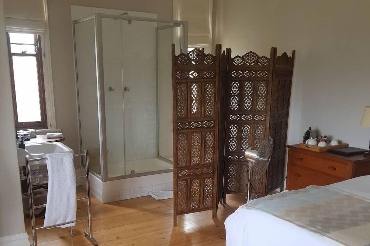 Un dormitorio con un armario de madera, una cama, una mesita de noche, un espejo y un baño con ducha y lavamanos.