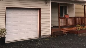 Ein zweistöckiges Haus mit Garage, einer Veranda mit Holzgeländer und einem kleinen Garten.