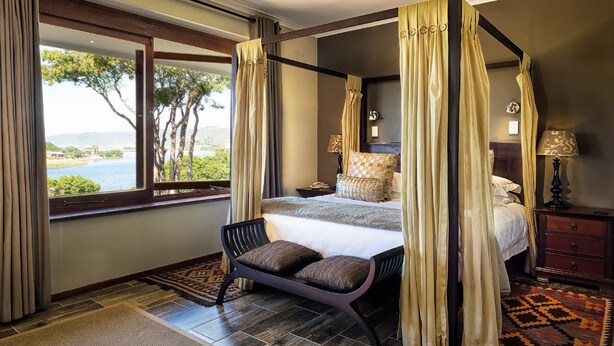 A four-poster bed with a view of the sea, a bench with cushions, and a patterned rug. - Wilderness Manor (Wilderness)