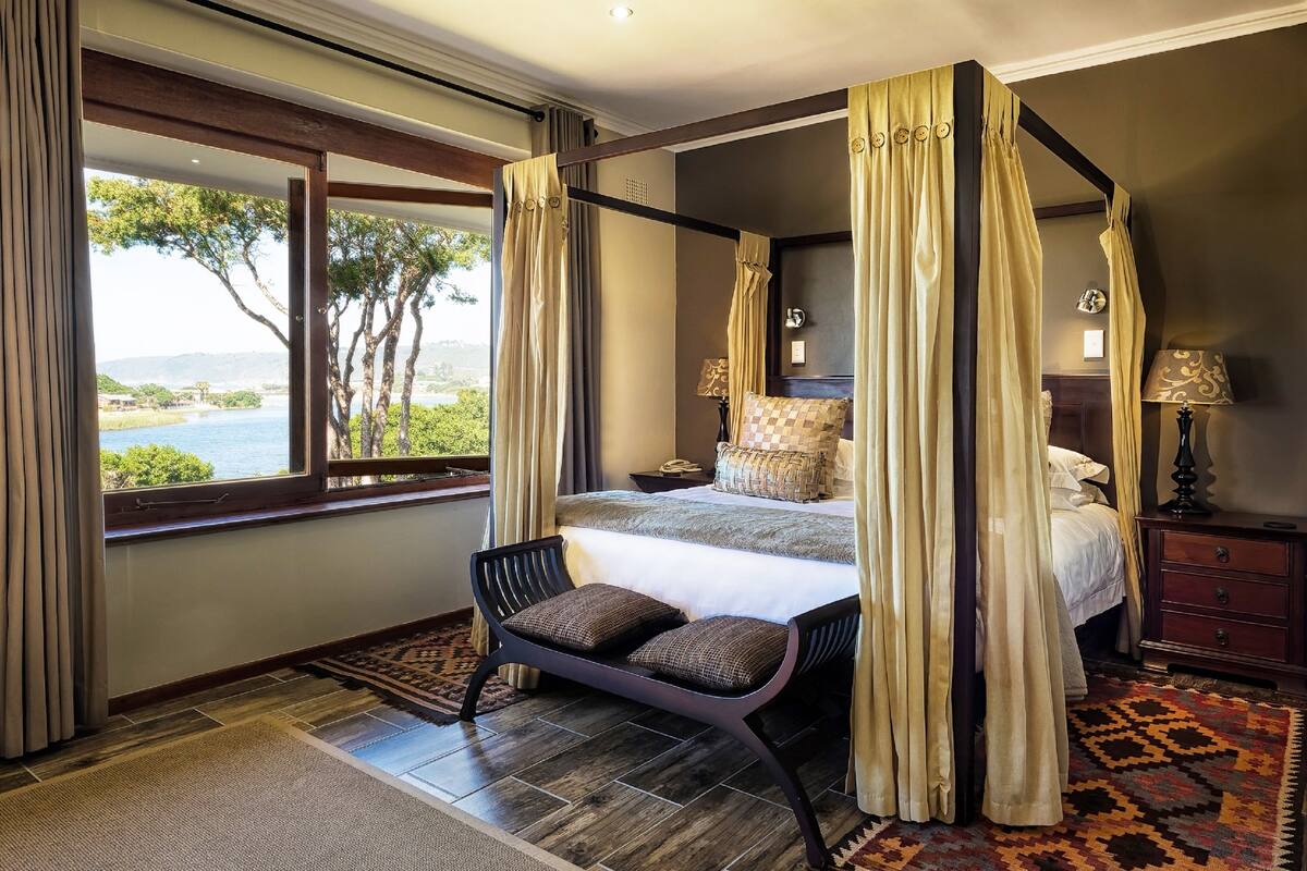 A four-poster bed with a view of the sea, a bench with cushions, and a patterned rug.