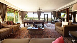A spacious living room with a piano, sofas, a coffee table, and large windows offering a view of the outdoors. - Wilderness Manor (Wilderness)