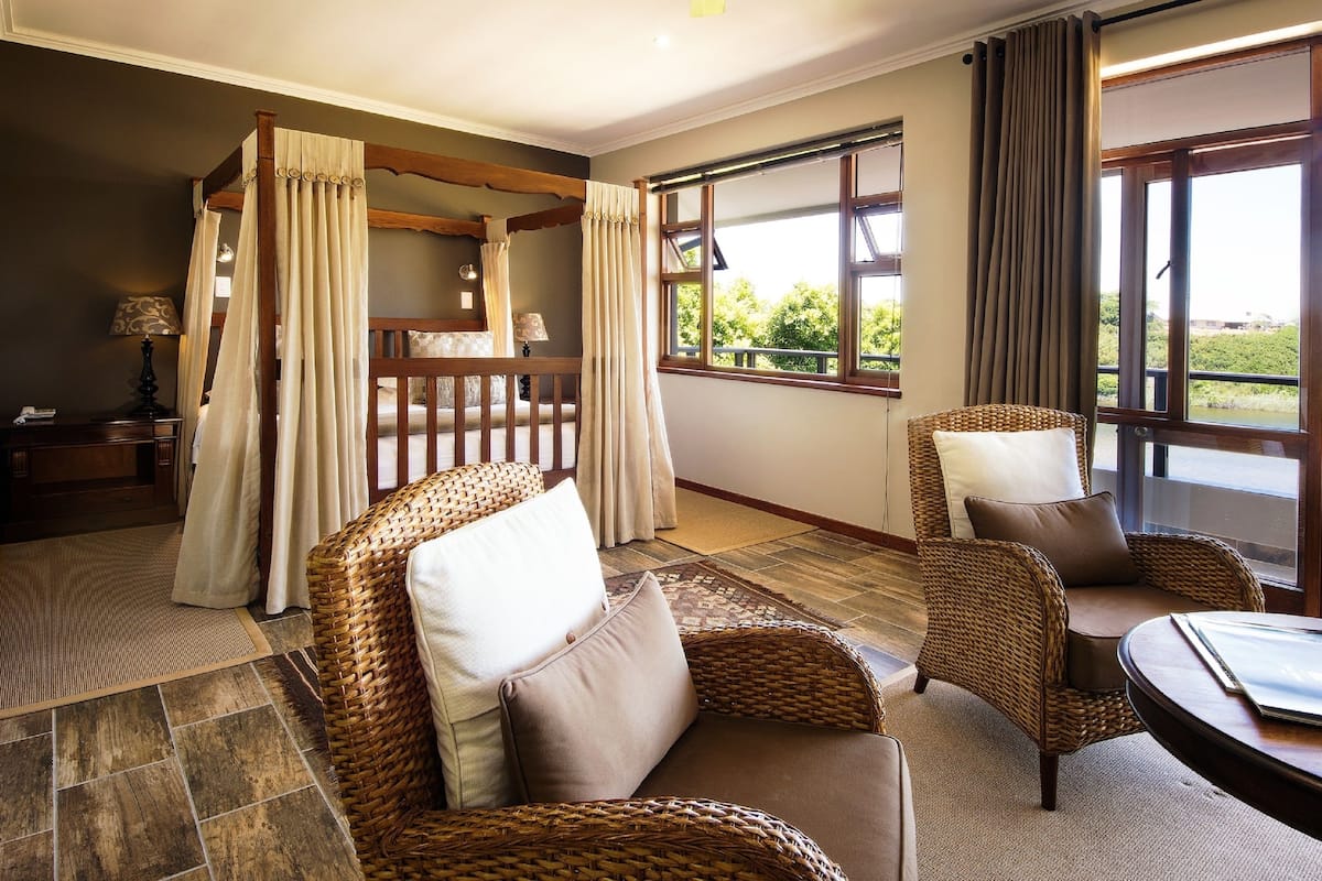 A room with a four-poster bed, wicker furniture, and large windows offering a view of greenery.
