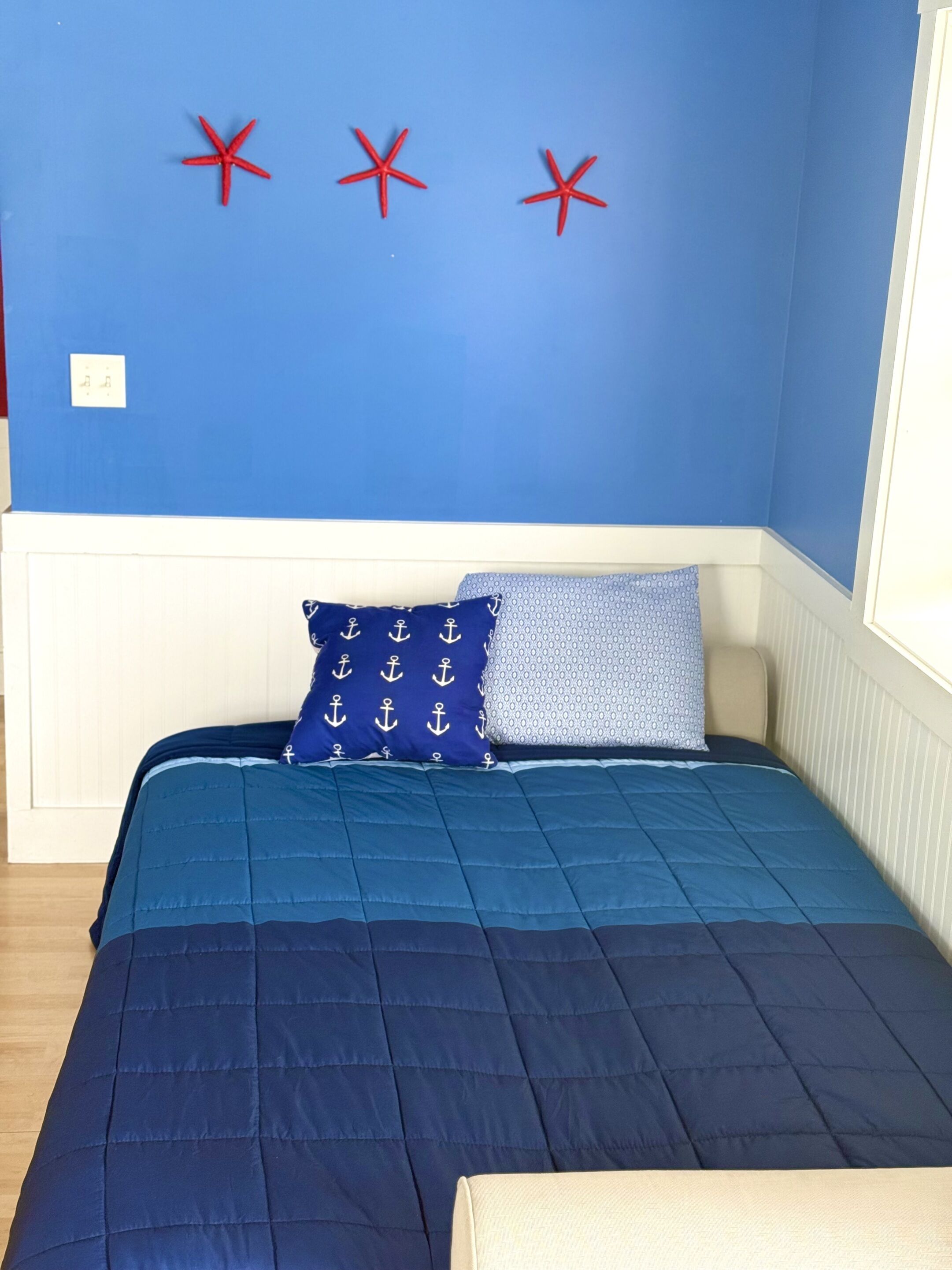 A bedroom with a blue wall, a bed with a blue quilt, two pillows with anchor patterns, and three red starfish decorations.