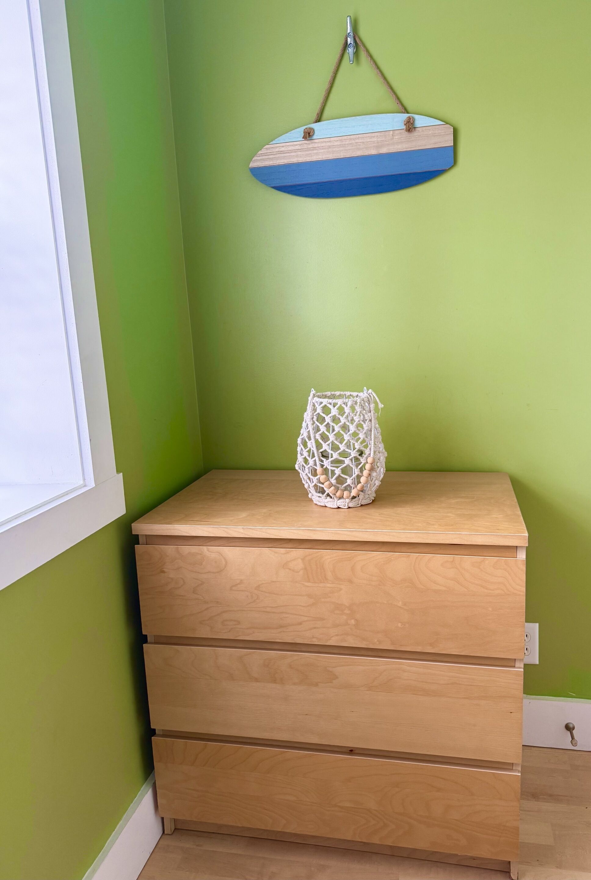 Un aparador de madera con tres cajones frente a una pared verde, un letrero de madera colgante y un adorno de macramé en la pared.