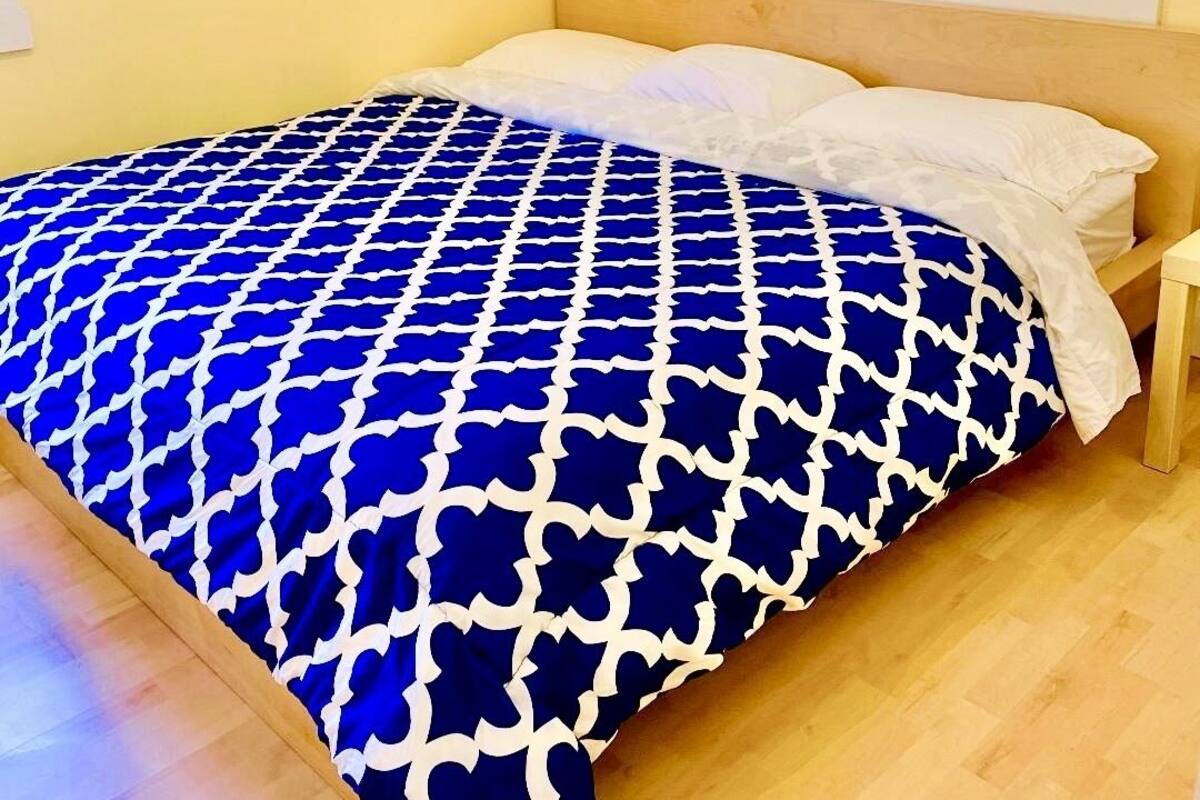 Ein ordentlich bezogenes Bett mit einer blau-weißen Bettdecke, einem Holz-Kopfteil und einem kleinen Topfpflanzen auf dem Fensterbrett.