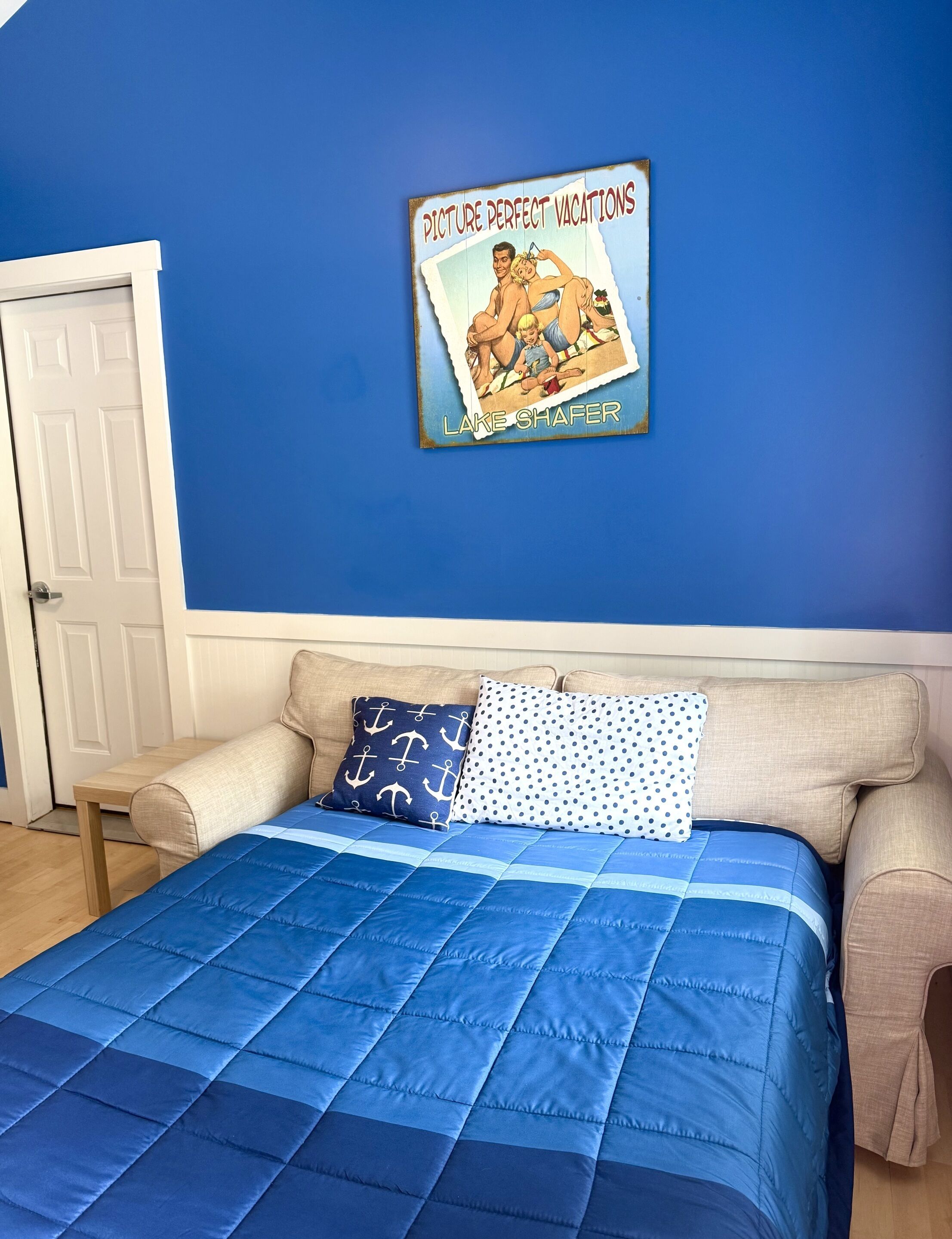 A bedroom with a blue wall, a bed with a blue quilt, and a poster of a vacation scene.