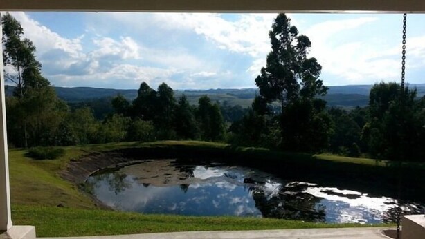 A view from a covered porch overlooking a pond with surrounding greenery and distant hills. - Arum Hill Lodge (Balgowan)