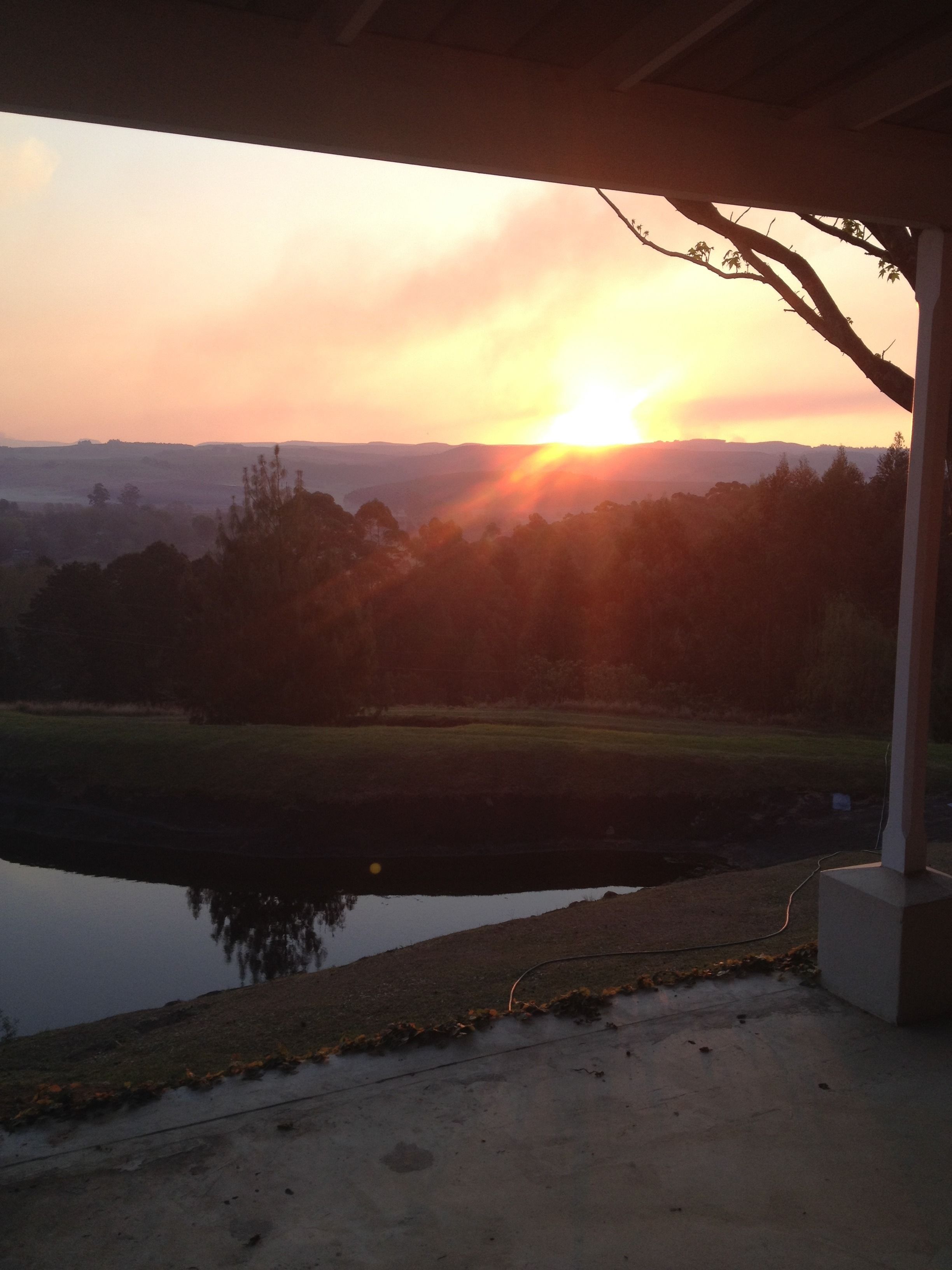 A sunset view from a covered outdoor area with a pond in the foreground.