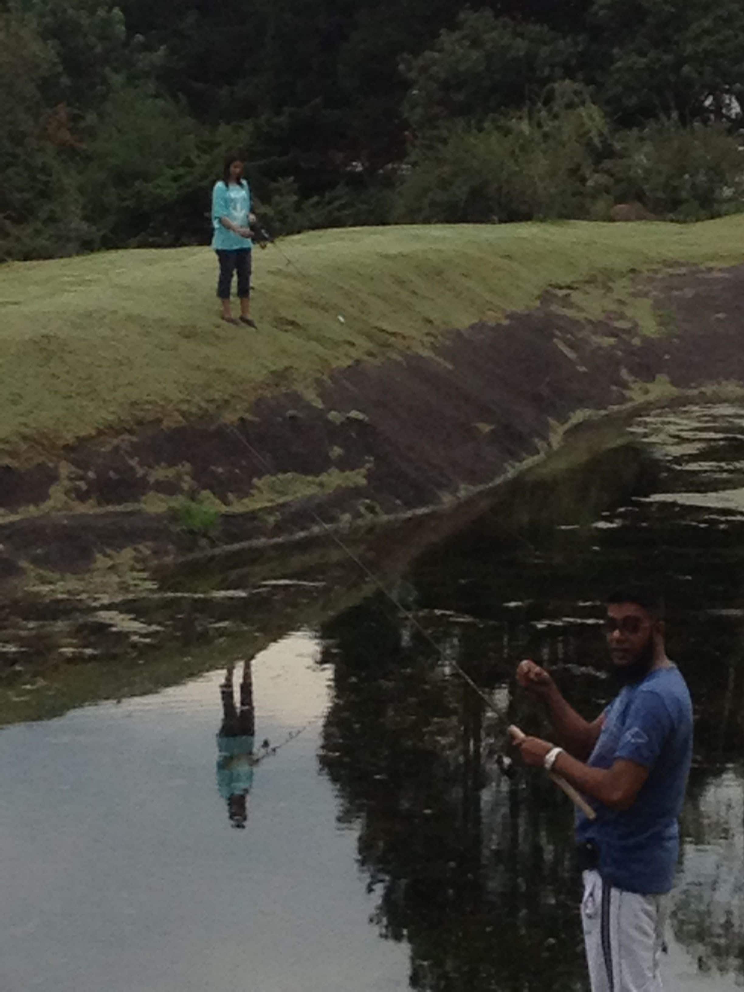 A person standing by a calm water body, possibly a pond or small lake, with a grassy area and trees in the background.