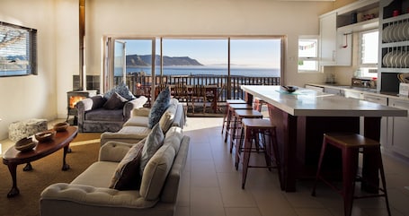 A living room with a sofa, a dining area with stools, a kitchen with a bar, and a large sliding glass door leading to a balcony with a view of the sea.. The Grosvenor Guest House