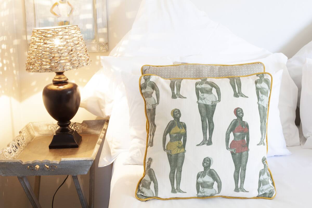 Ein Bett mit weißen Leinen, ein Dekokissen mit einem Muster von menschlichen Figuren und eine Tischlampe auf einem hölzernen Nachttisch.