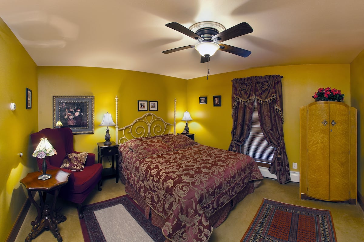 A bedroom with a bed, a red armchair, a nightstand with a lamp, a ceiling fan, a wardrobe, and a window with curtains.