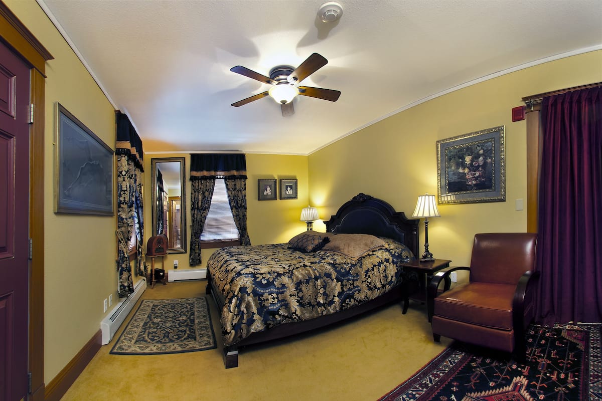 A bedroom with a bed, a chair, a ceiling fan, a nightstand, a lamp, and a rug.