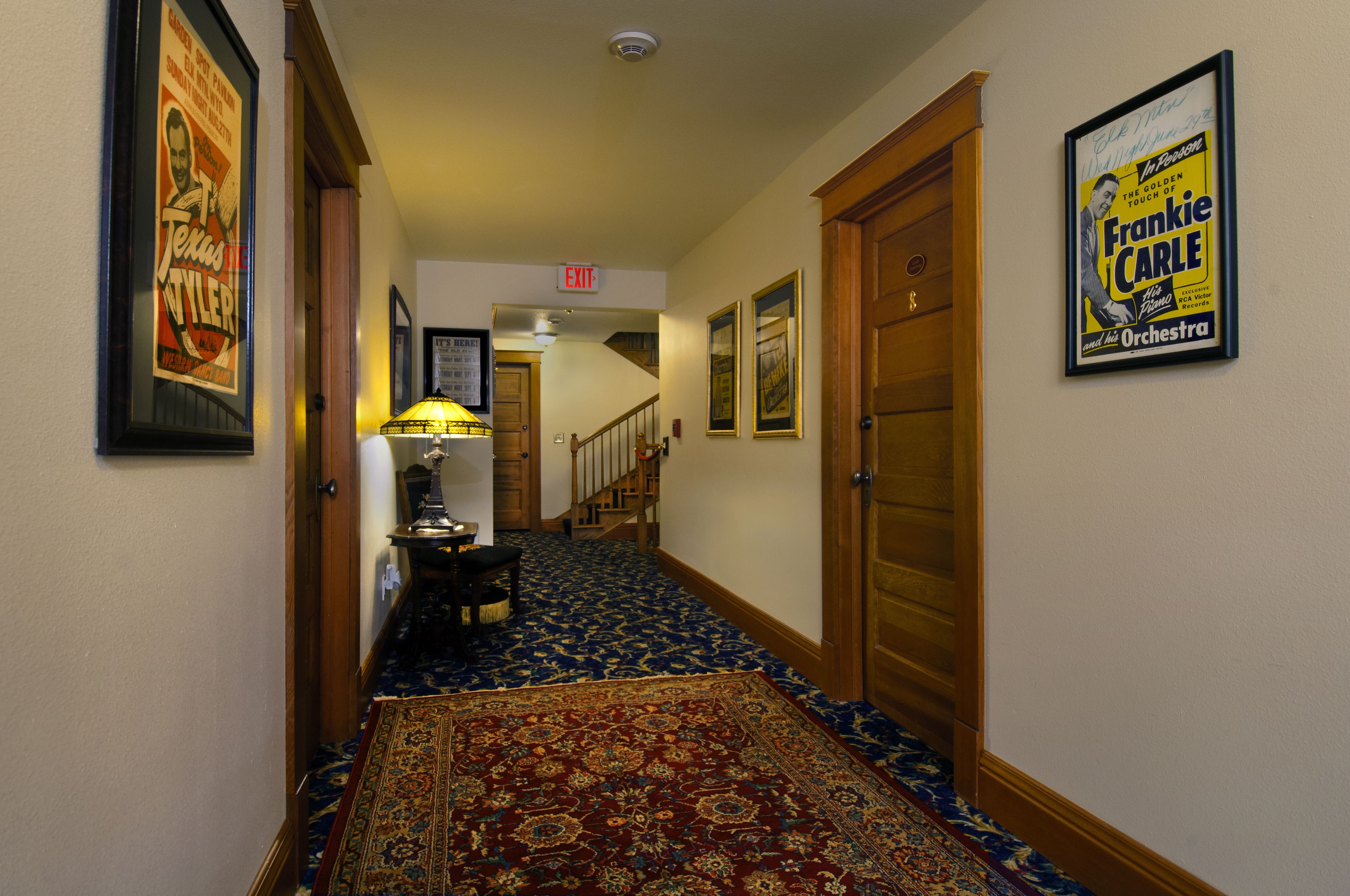 A hallway with a patterned carpet, framed pictures on the walls, and a framed poster of Frankie Carle.