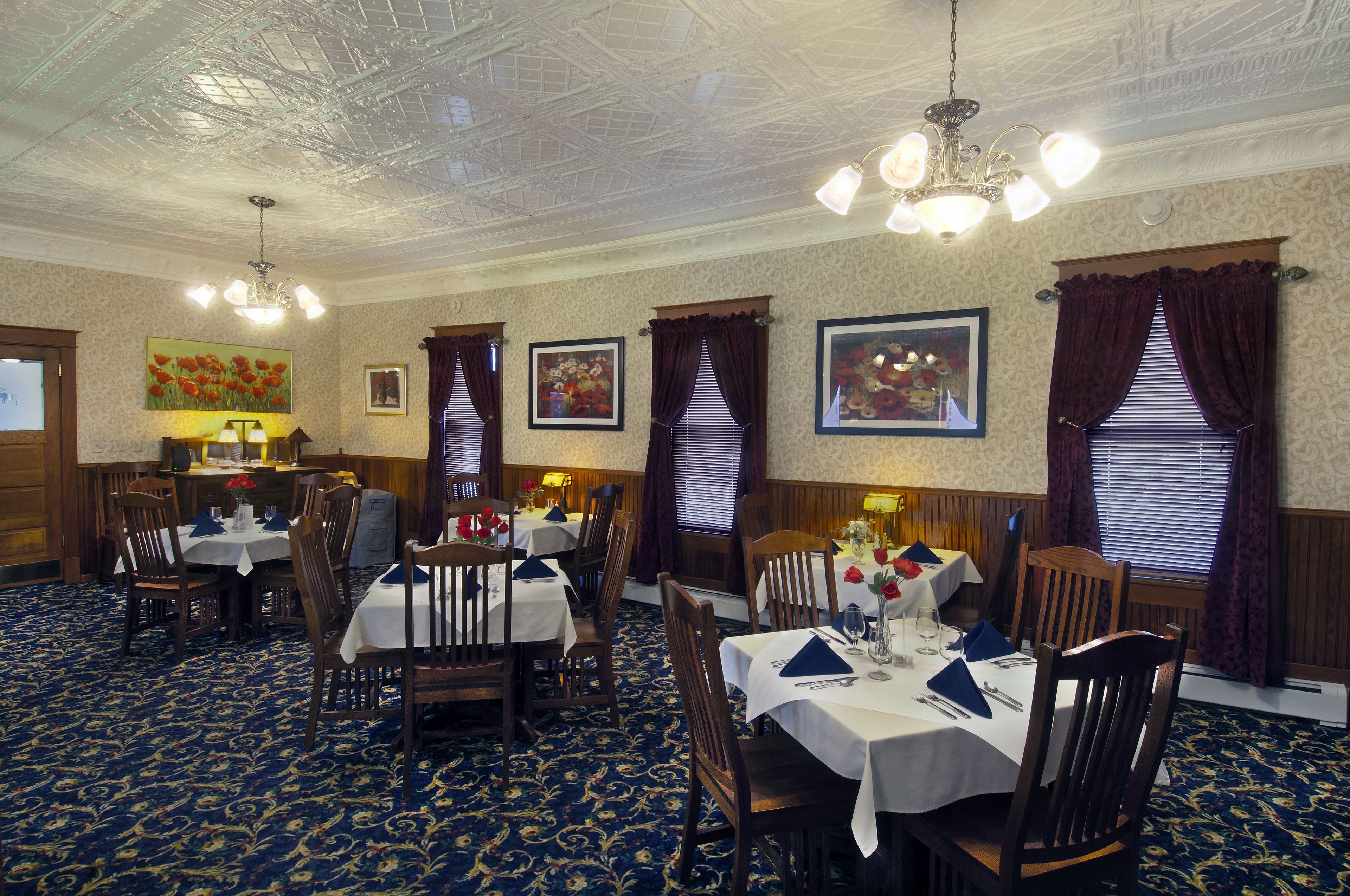 Une salle à manger avec plusieurs tables dressées pour les convives, agrémentée d’un lustre classique, d’un papier peint à motifs et d’œuvres d’art encadrées qui ornent les murs.