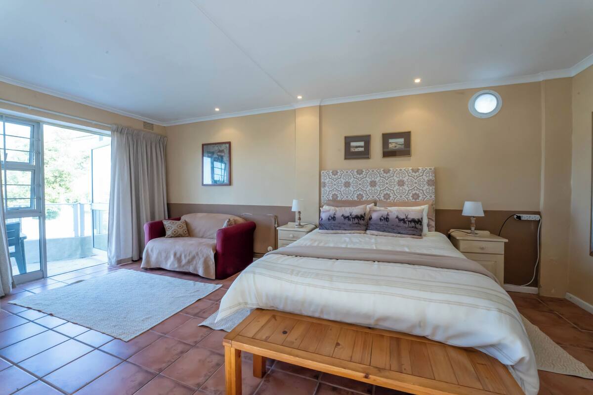 A bedroom with a large bed, a red chair, a bedside table with a lamp, and a window with curtains.