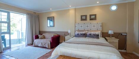 Un dormitorio con una cama grande, una silla roja, una mesita de noche con lámpara y una ventana con cortinas.