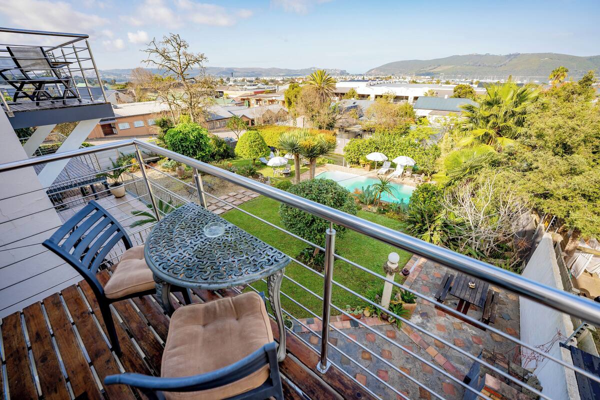 A balcony with a table and chairs, a pool, and a view of a residential area with houses and hills.
