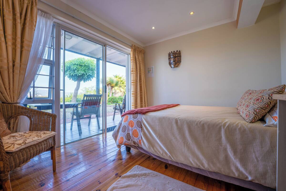 A bedroom with a bed, a wicker chair, a glass door leading to an outdoor area, and a wall-mounted decoration.
