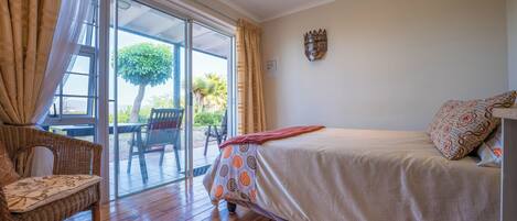 Un dormitorio con una cama, una silla de mimbre, un ventanal de vidrio que da a un espacio exterior y una decoración en la pared.