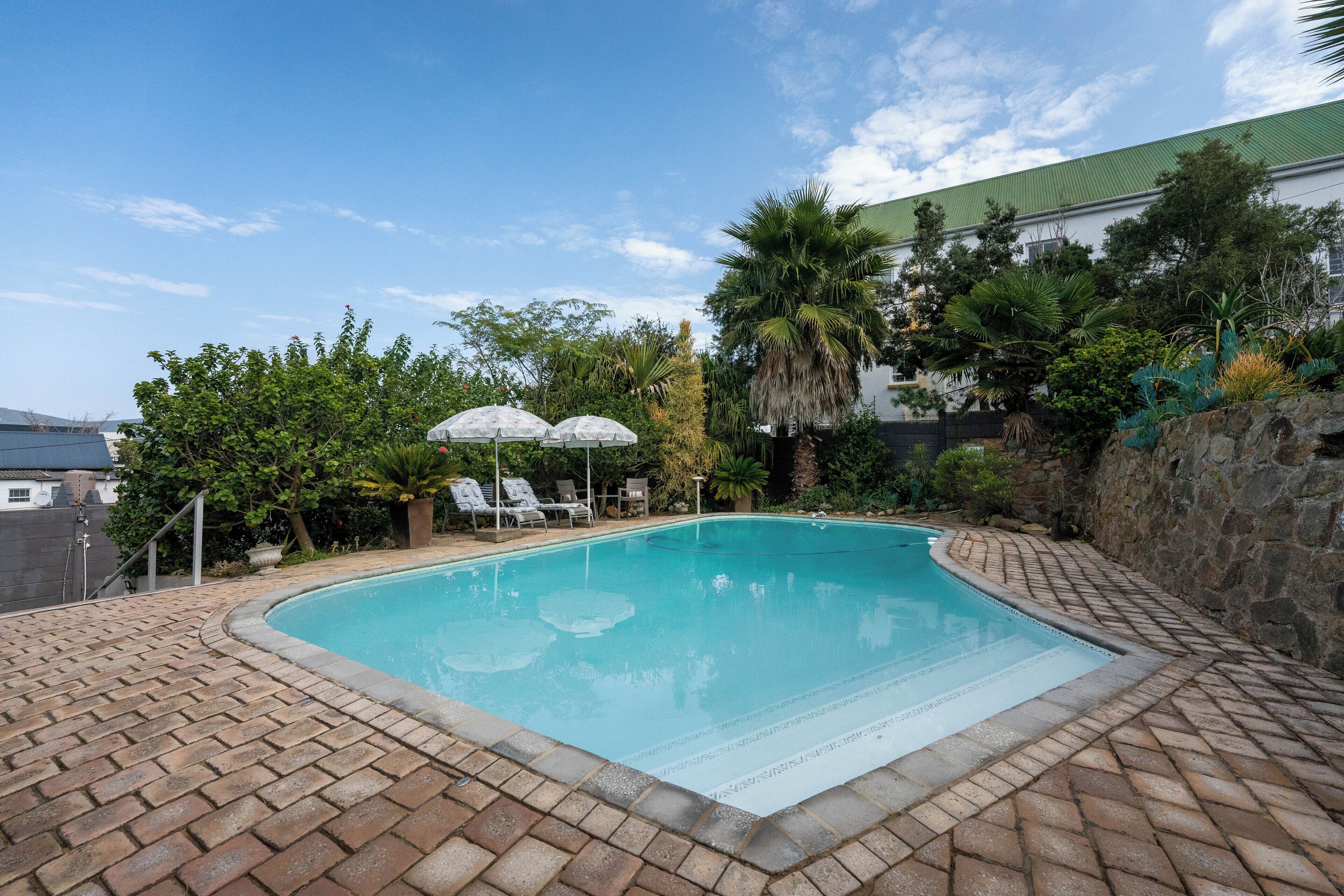 A swimming pool with a brick border, surrounded by lounge chairs and umbrellas, with a stone wall and greenery in the background.