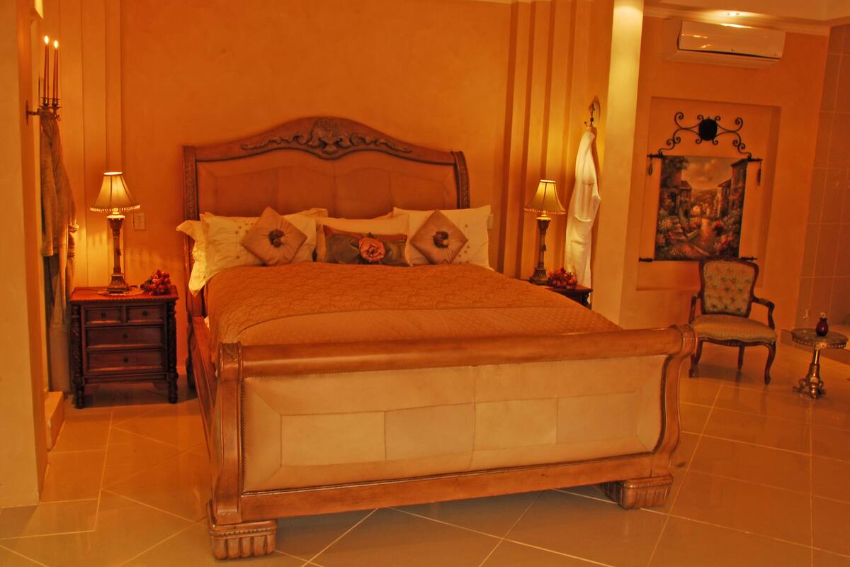A large bed with a wooden headboard, two bedside tables with lamps, a chair, and a painting on the wall.