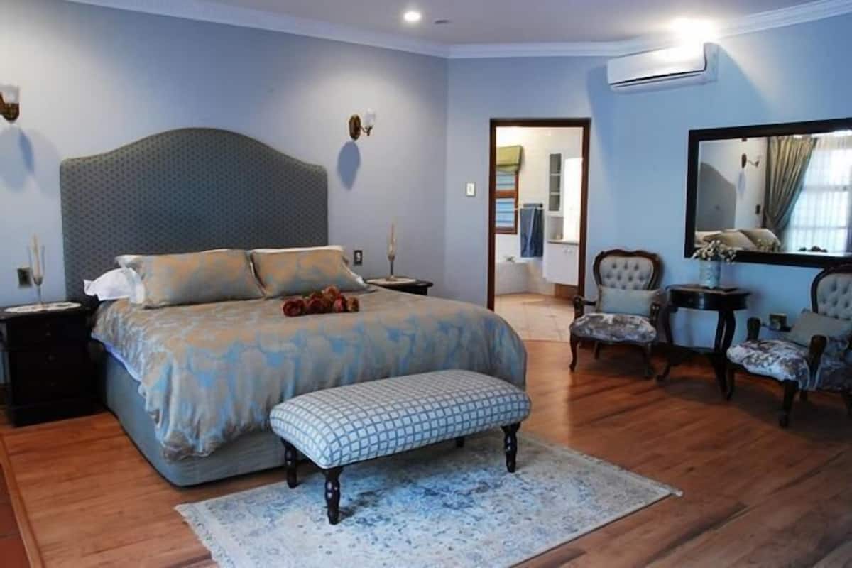 A bedroom with a large bed, a bench, a chair, a small table, and a mirror.