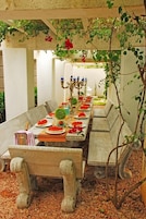 An outdoor dining area with a long table set for a meal, surrounded by stone benches and a pergola with climbing plants.