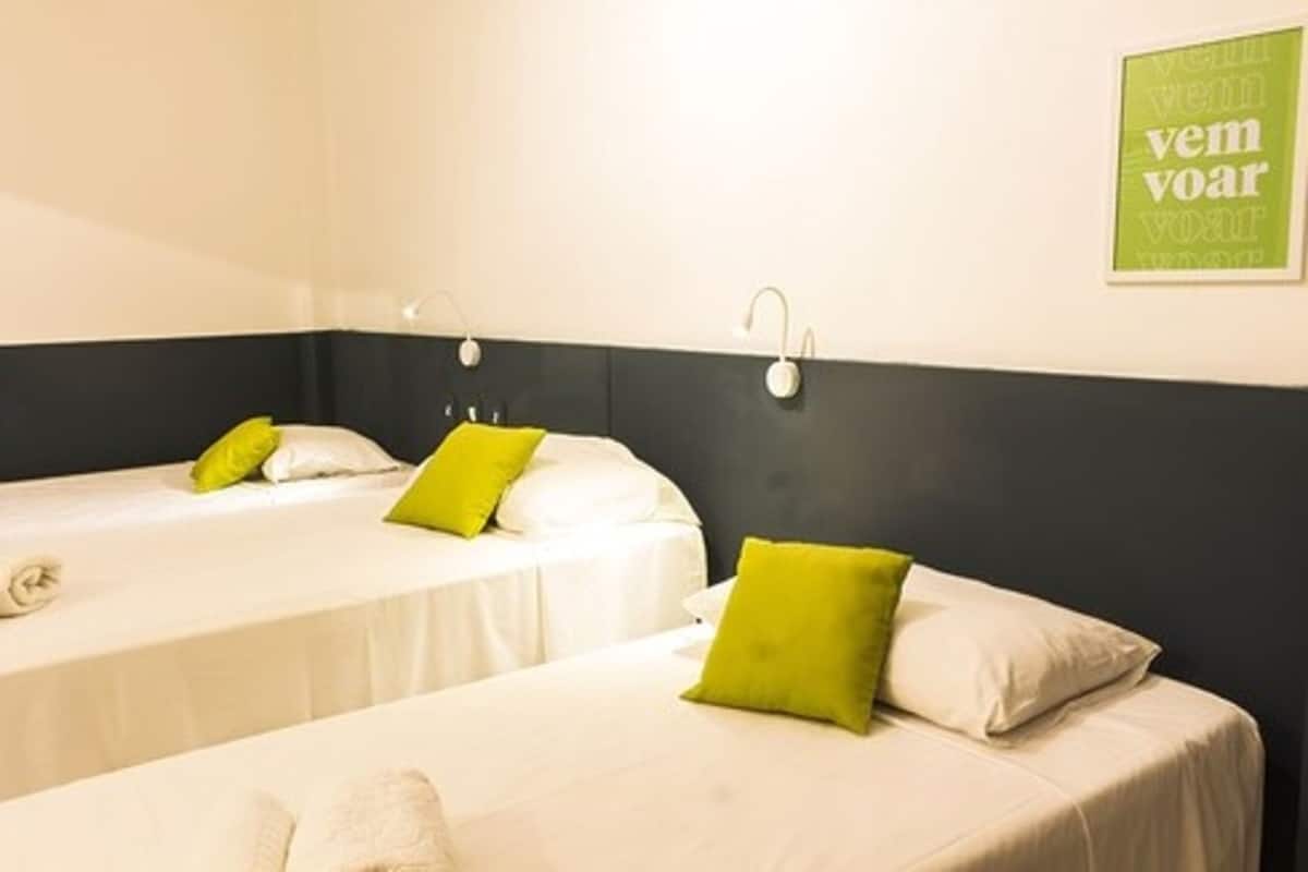 Une chambre d’hôtel avec deux lits, chacun étant équipé de draps blancs et d’oreillers verts. Des lampes murales sont fixées au-dessus de chaque lit.