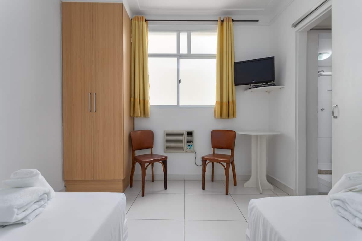 Une chambre d’hôtel avec une armoire en bois, deux chaises en bois, une petite table blanche, un téléviseur fixé au mur et un espace douche.
