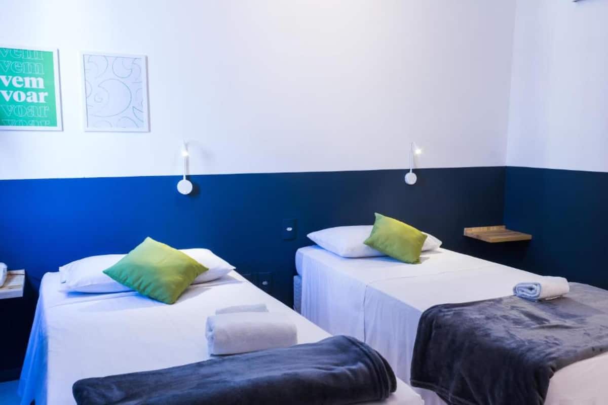 Une chambre d’hôtel avec deux lits, chacun équipé d’un oreiller et d’une serviette. On y trouve des lampes fixées au mur et une petite étagère en bois.