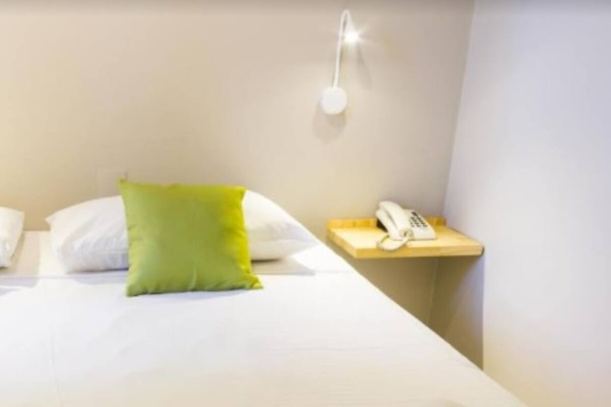 Un lit avec une literie blanche et deux oreillers verts, une table de chevet avec un téléphone et deux lampes fixées au mur.