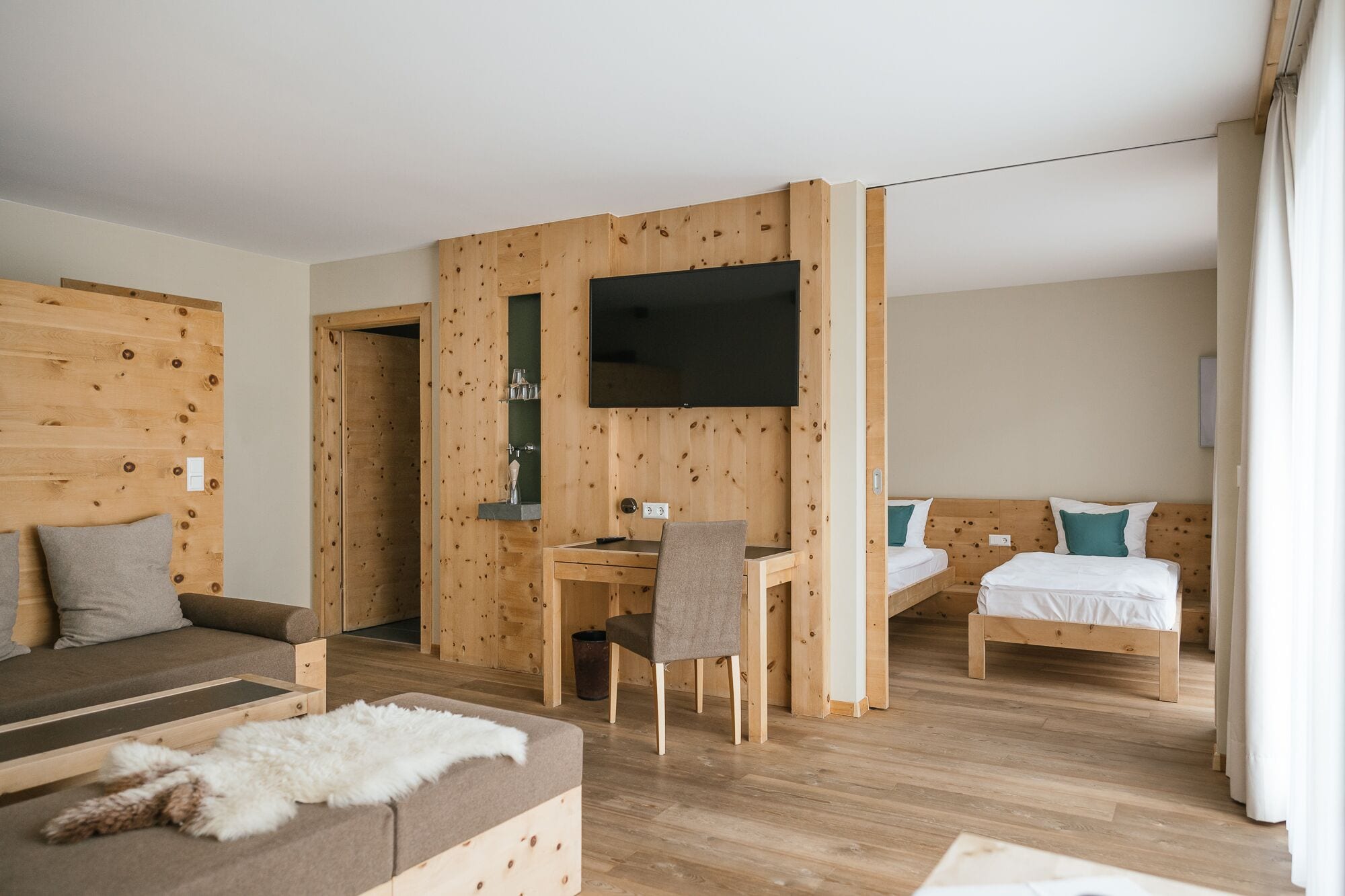 A hotel room with a wooden wall, a flat-screen TV, a desk with a chair, two beds, and a sofa.