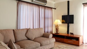 Een woonkamer met een beige bank, een houten dressoir, een flatscreen televisie en een lamp.