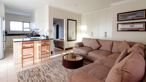 Een moderne woonkamer met een beige bank, een salontafel, en een keukenhoek met witte kasten en een barkruk.
