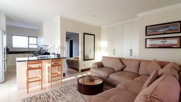 Una sala moderna con un sofá color beige, una mesa de centro y una zona de cocina con armarios blancos y un taburete de bar.