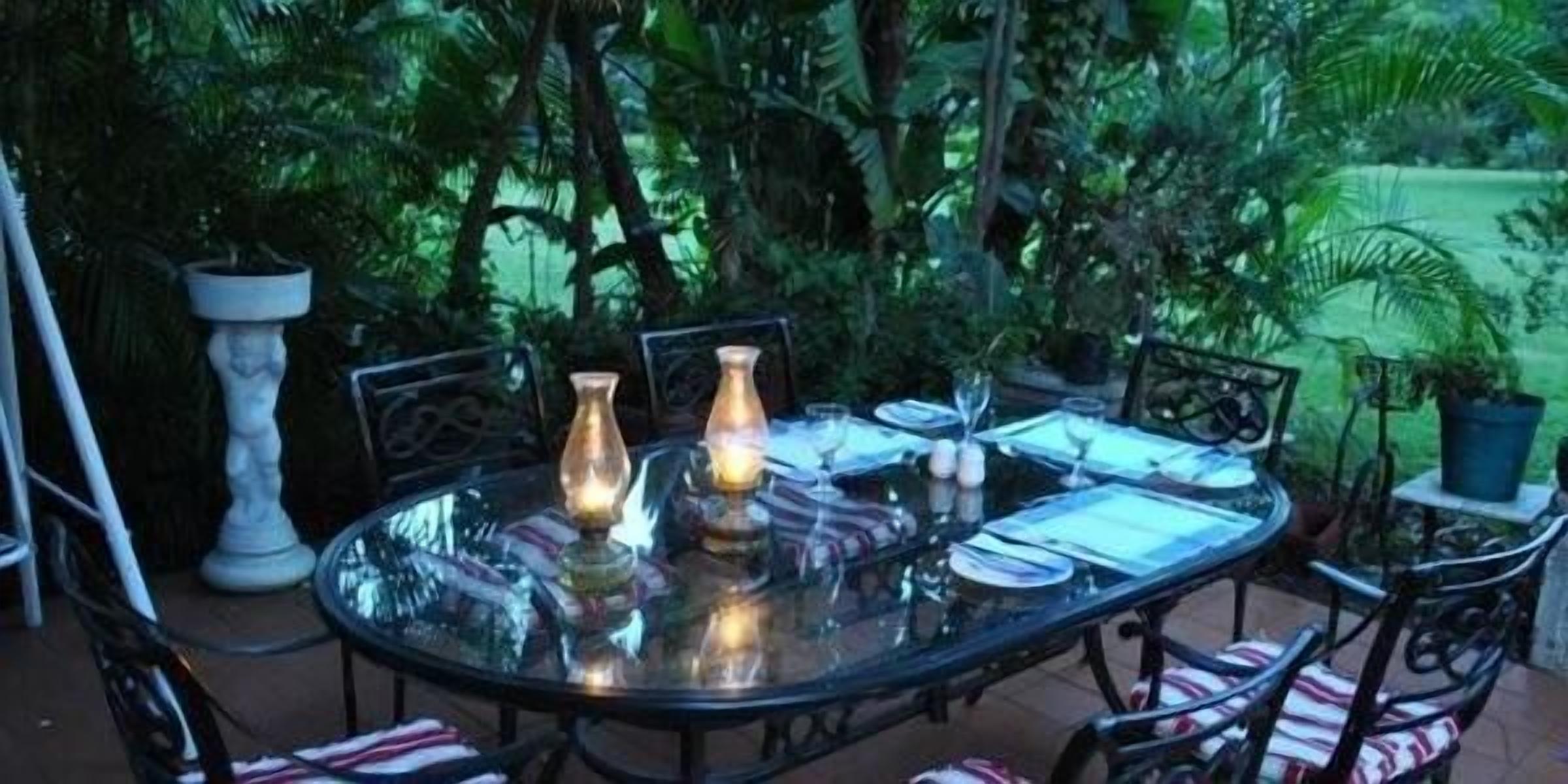 Mesa con cubierta de vidrio lista para comer al aire libre, con sillas, rodeada de abundante follaje.