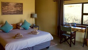 A bedroom with a bed, pillows, a nightstand, a lamp, a desk with a chair, and a window with curtains.