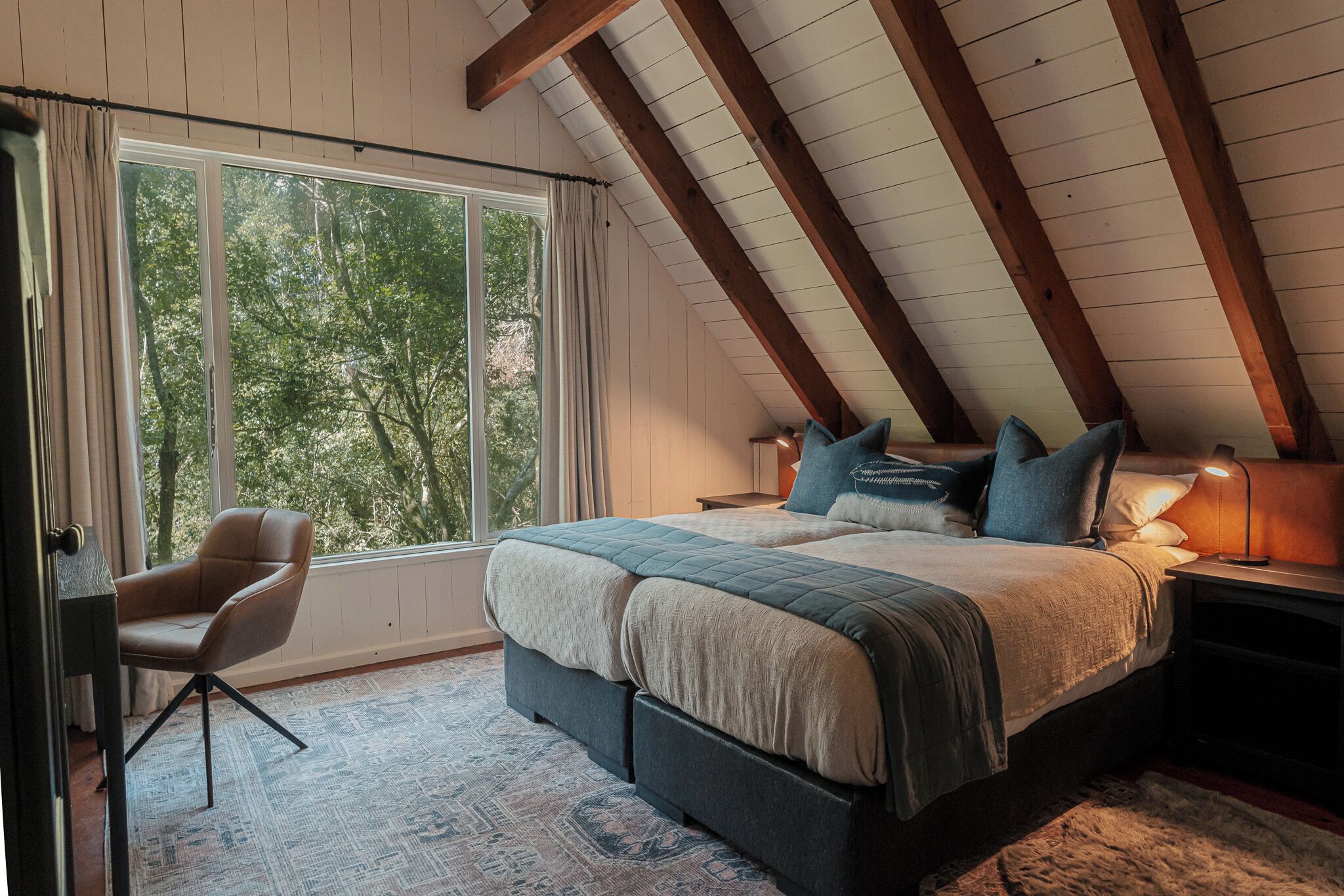 A bedroom with a large window, a bed with pillows, a chair, and a bedside table.
