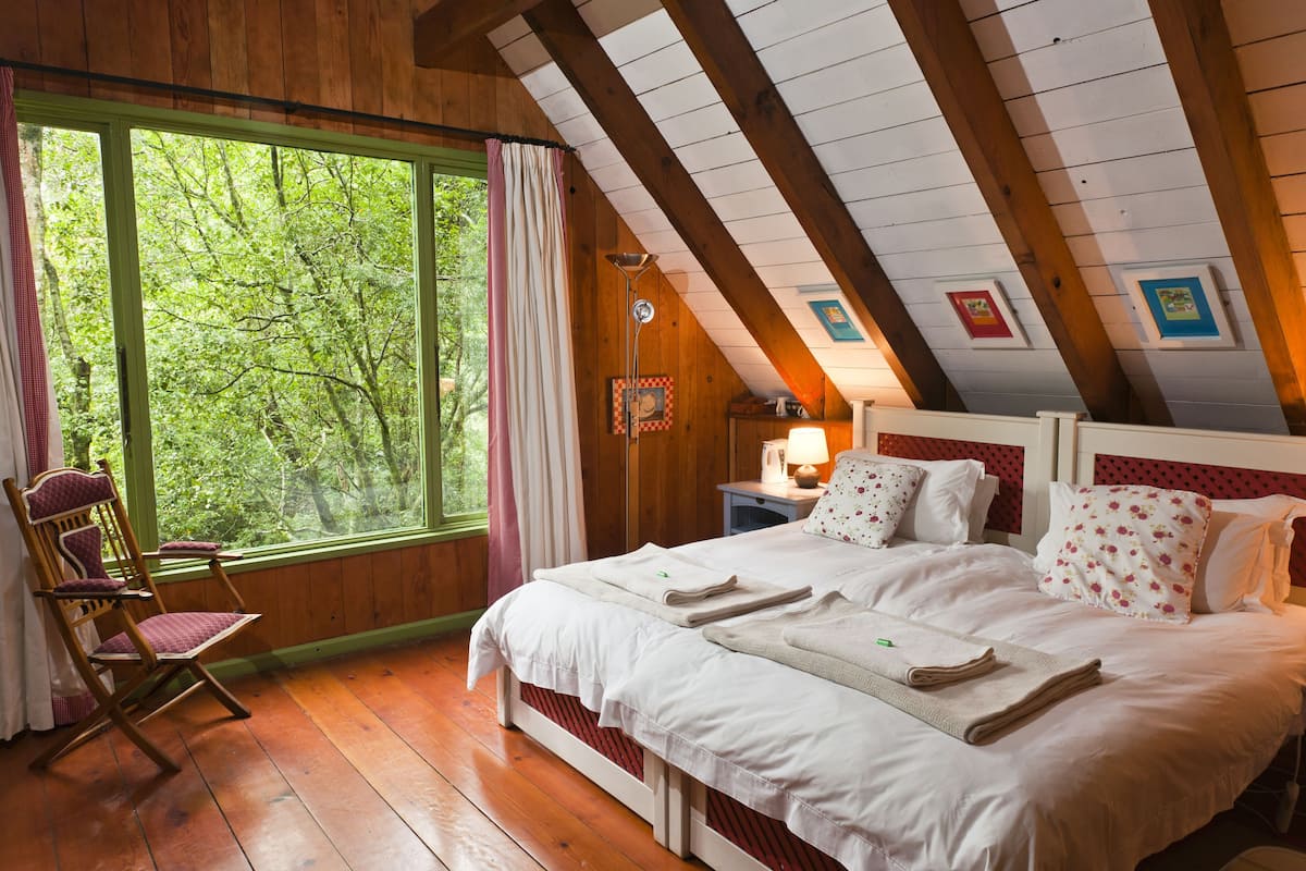 A bedroom with a large window, wooden beams, a bed with white linens, and a chair.