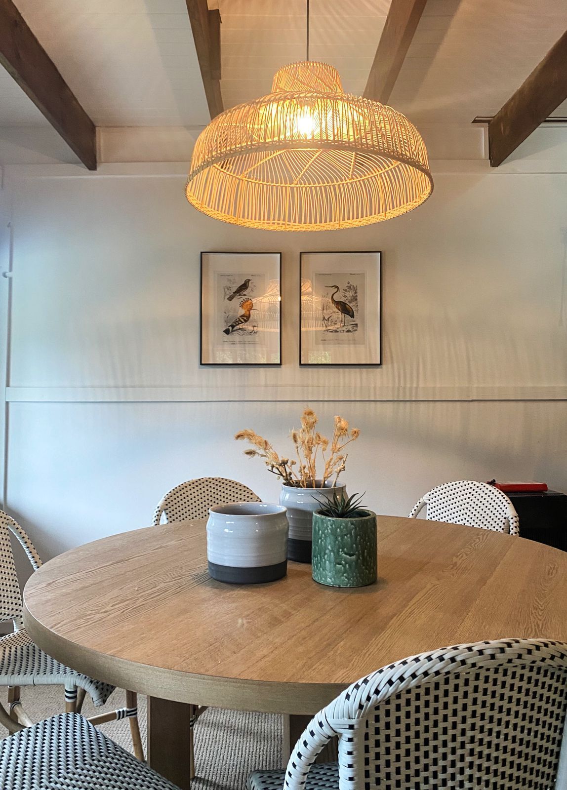 Una mesa de comedor redonda de madera con una lámpara colgante trenzada encima, rodeada de sillas de mimbre.