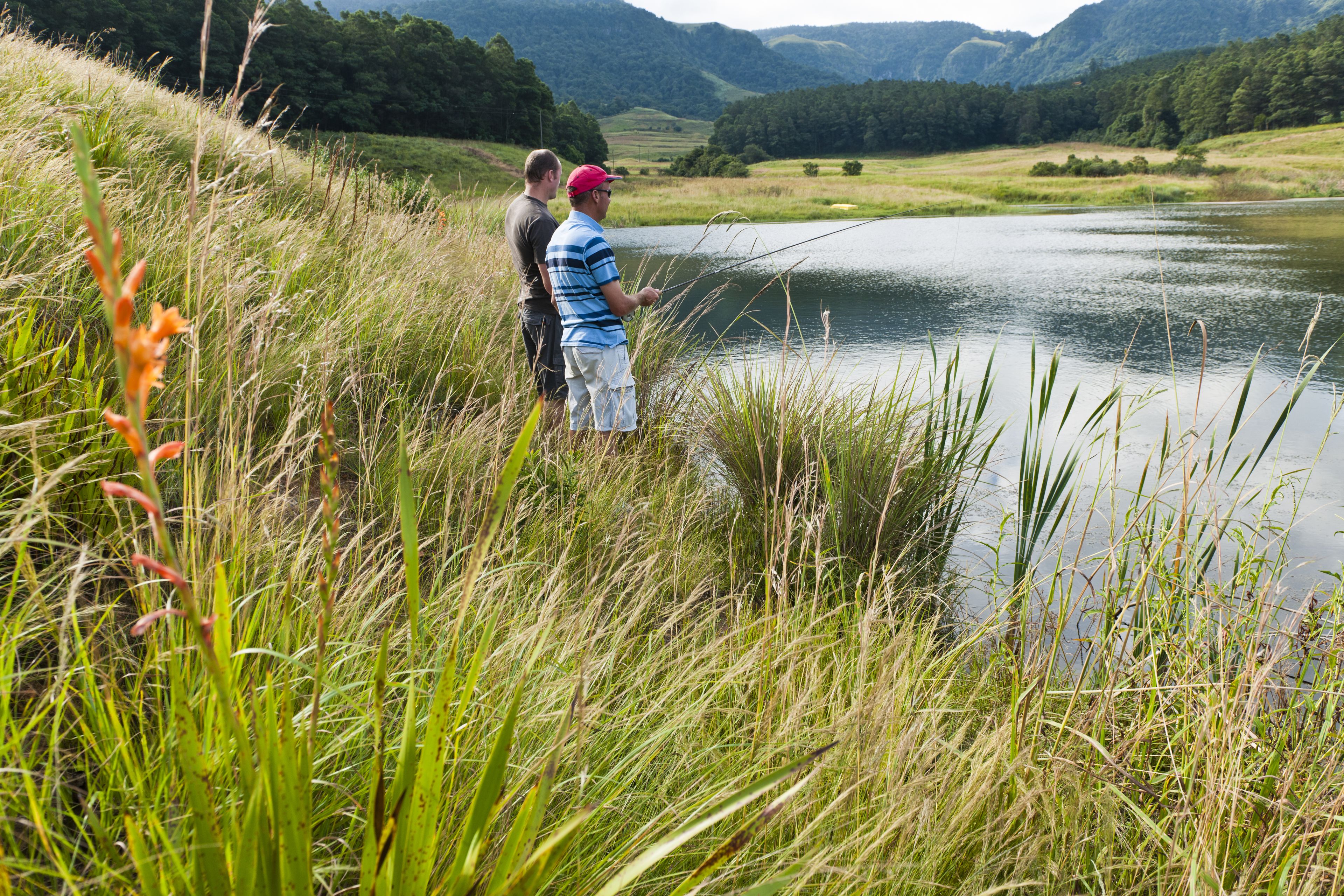 Dos personas al lado de un lago tranquilo rodeado de hierba alta y montañas al fondo.