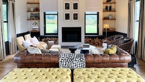 A spacious living room with a large leather sofa, a patterned throw pillow, and a fireplace. There are shelves with decorative items and a view of the outdoors through large windows. - Rockwood Lodges (Karkloof Nature Reserve)