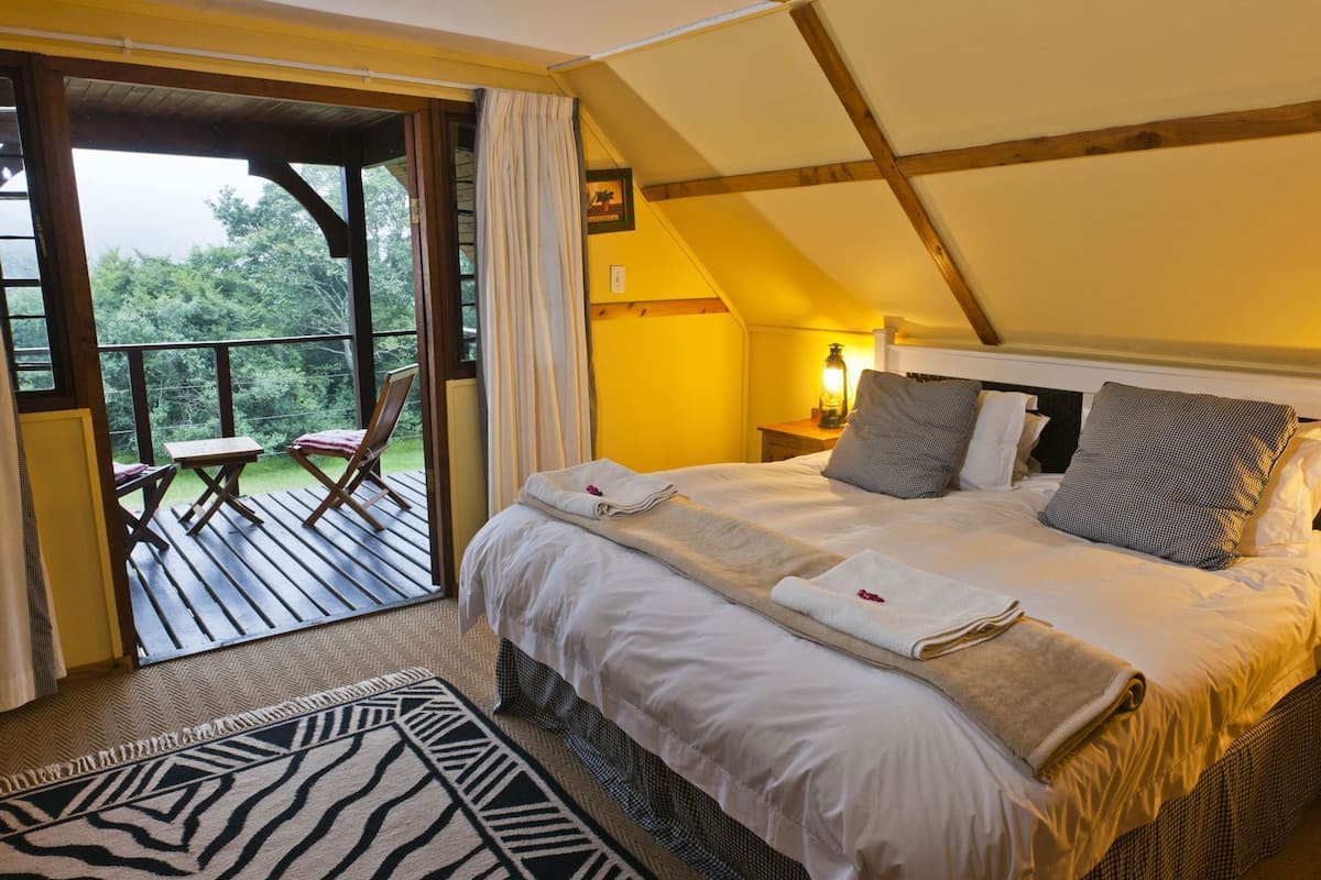 A bedroom with a bed, pillows, a nightstand, a lamp, a window, and an outdoor deck with chairs.