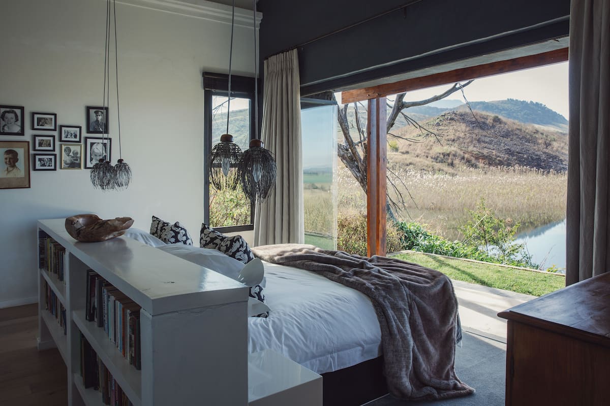A bedroom with a large window, a bookshelf, and a view of a lake and hills.