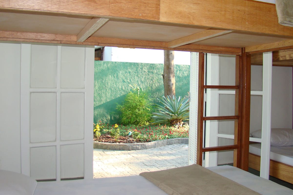 A bedroom with a wooden ceiling, a bed, and a view of a garden with plants and a tree.