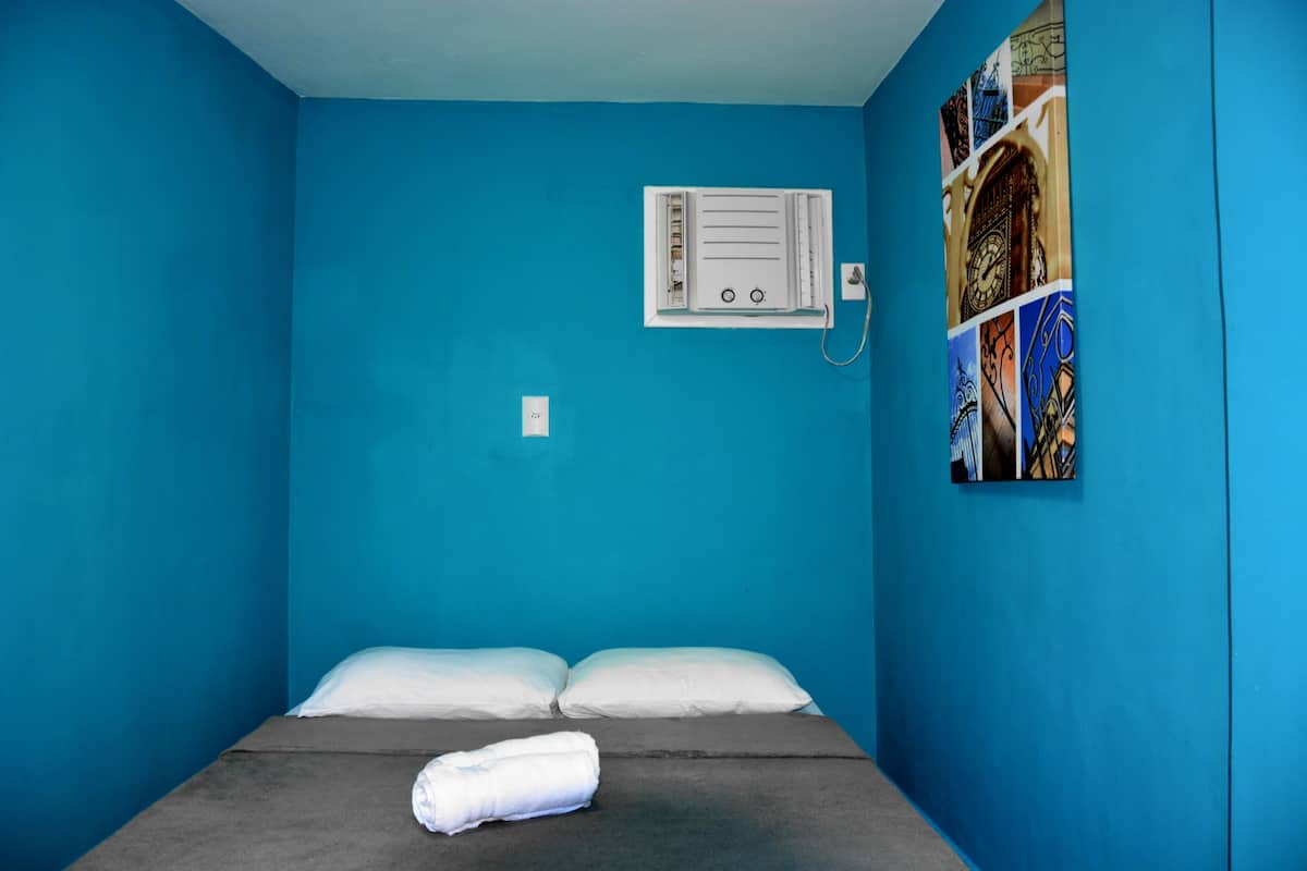 A small, single-bed room with a blue wall, a white air conditioning unit, and a wall-mounted artwork.