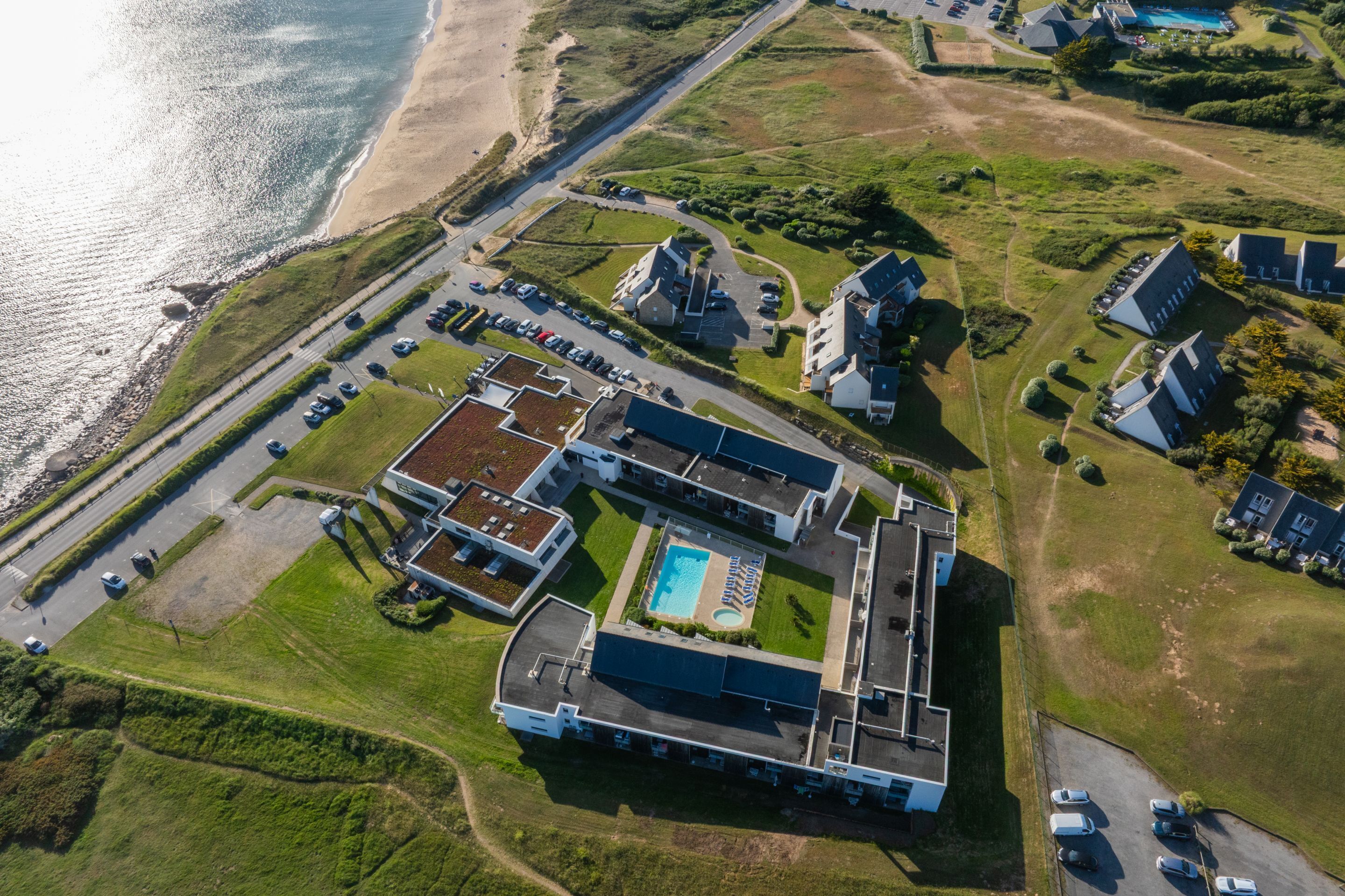 Vue aérienne d’un hôtel situé sur la côte, composé de plusieurs bâtiments, avec une piscine et un parking.