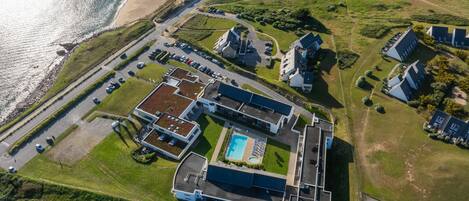 Vue aérienne d’un hôtel situé sur la côte, composé de plusieurs bâtiments, avec une piscine et un parking.